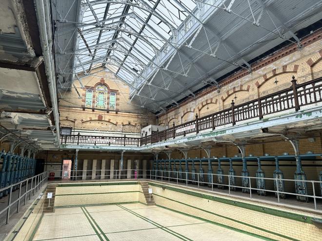 The empty, tiled Gala Pool at Victoria Baths beneath its glazed roof, ringed by tiered changing cubicles